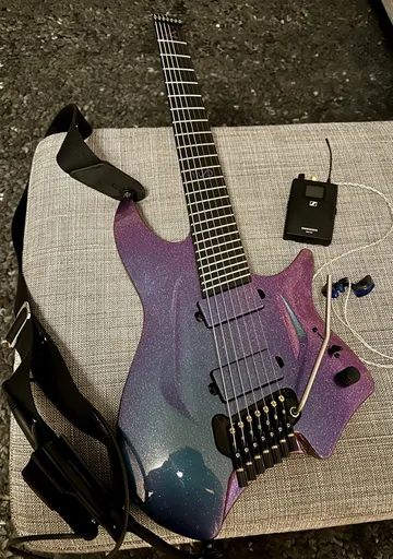 Aristides H/07 headless seven-string guitar with a purple-to-teal chameleon finish under silver sparkle and matching painted pickups, lying on a gray cushion.